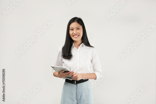 Photo of an Asian woman 20s standing and holding a tablet, dressed a white shirt and isolated on a white background.