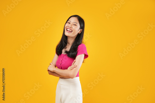 An Asian woman crosses her arms and smiles warmly, displaying confidence and approachability against a bright yellow background.