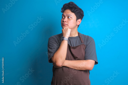 Asian barista man in brown apron touching his chin and looking to the side over blue background