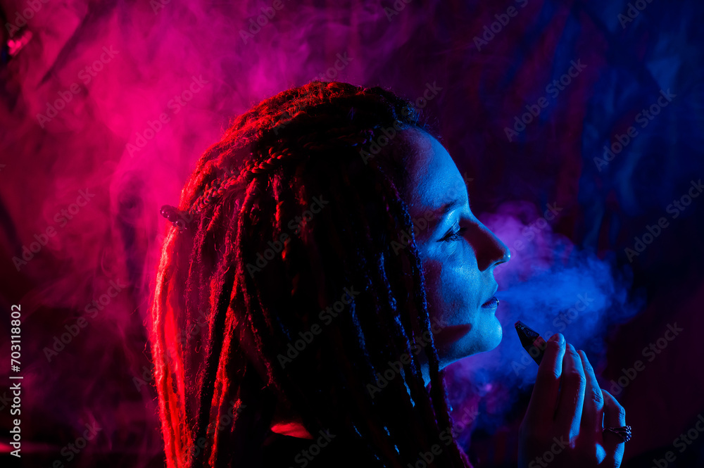 Profile of caucasian girl with dreadlocks smokes a vape in red blue light. 