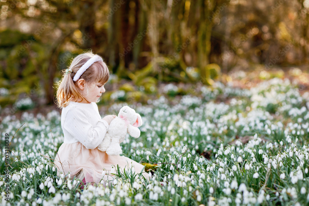 Adorable little girl with Easter bunny ears holding soft plush toy in spring forest on sunny day, outdoors. Cute happy child with lots of snowdrop flowers. Springtime, christian holiday concept.