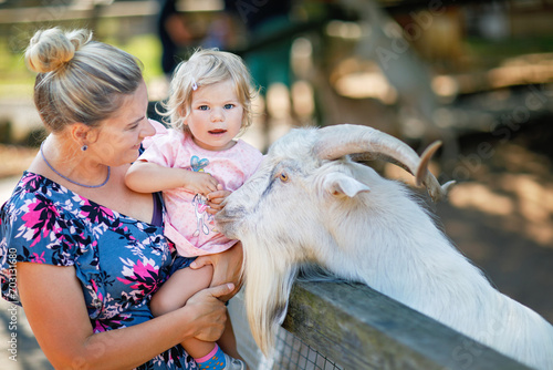 Adorable cute toddler girl and young mother feeding little goats and sheeps on kids farm. Beautiful baby child petting animals in petting zoo. Woman and daughter together on family weekend vacations © Irina Schmidt