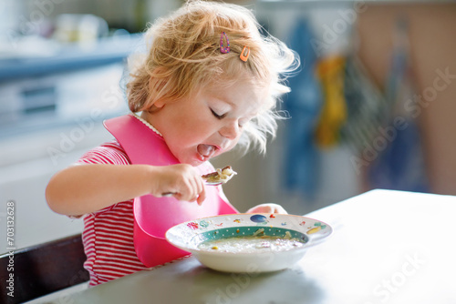 Adorable baby girl eating from spoon vegetable noodle soup. Healthy food, child, feeding and development concept. Cute toddler child with spoon sitting in highchair and learning to eat by itself photo
