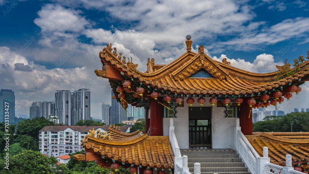 Obraz premium A Chinese Thean Hou Temple against a blue sky, clouds, city skyscrapers. The elegantly curved roof is decorated along the edge with row of red traditional lanterns. Stairs with white carved handrails