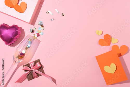 A chocolate bar, paper hearts, blank card, a heart balloon and a clear wine glass on a pink background. Empty space with a flat top-down view.