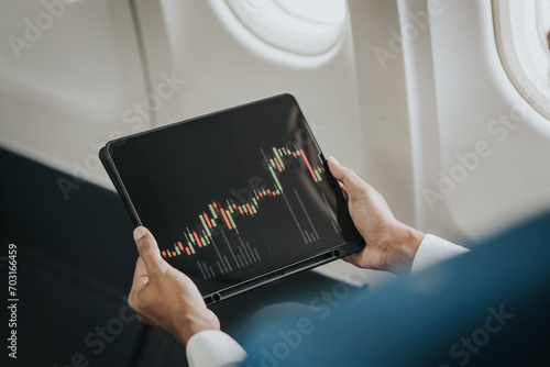 Fotografia Asian businessman, possibly a stock market trader or investor, focused on his tablet while flying, indicative of a professional managing work on the go