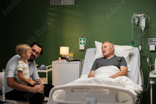 A grandson visits his grandfather in the hospital. The elderly man is lying in a hospital bed and is very happy.