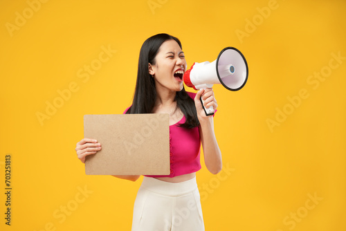 Portrait of a young Asian woman shouting angrily using a megaphone and holding paper. Isolated on a yellow background. Demonstration concept.