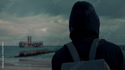 Young male in coat standing on the top of the hill and looking at the horizon with ruined building in the distance backview. Concept of solitude to non tourist spots solitude.
