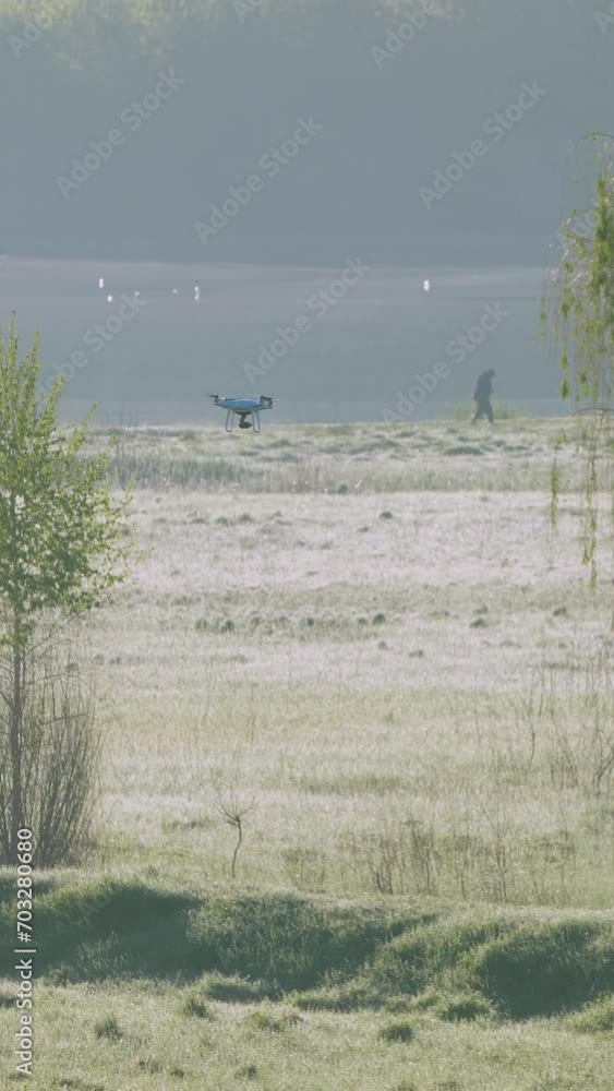 Vertical video. A white quadcopter in flight over a summer landscape. Morning dew and fog near the river, a person walks along the shore. A drone follows the man. Vertical video. A white quadcopter in flight over a summer landscape. Morning dew and fog near the river, a person walks along the shore. A drone follows the man.