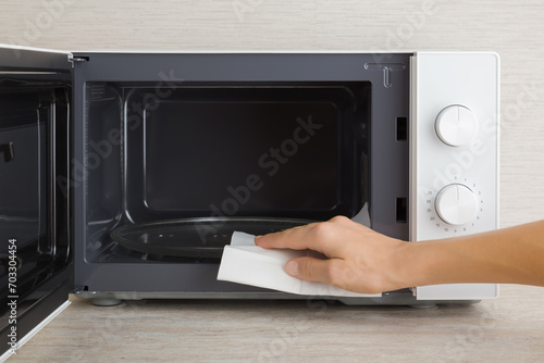 Young adult woman hand holding dry white paper napkin and wiping dark black surface of white microwave oven inside on table top at home kitchen. Closeup. Regular cleanup. Front view.