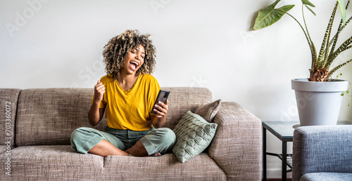 Excited happy young black woman holding smart phone device sitting on sofa at home - Happy satisfied female looking at mobile smartphone screen gesturing yes with clenched fist - Technology concept photo
