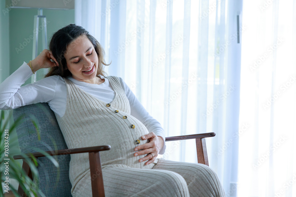 Happy pregnant young woman touching her pregnant belly and sitting near the window Happy pregnant young woman touching her pregnant belly and sitting near the window