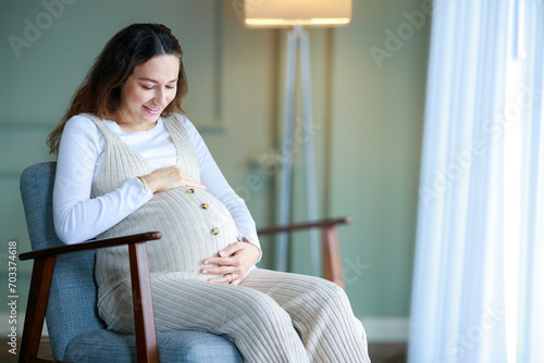 Happy pregnant young woman touching her pregnant belly and sitting near the window