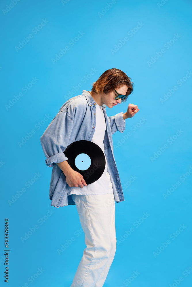Fototapeta premium Side view portrait of happy, overjoyed man, student in modern sunglasses dancing with vinyl record against blue studio background. Concept of retro music and dance, self-expression, party, fashion.