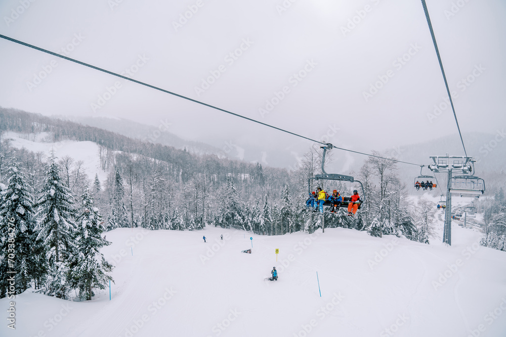 Tourists ride on a chairlift above the forest and ski slope with skiers skiing