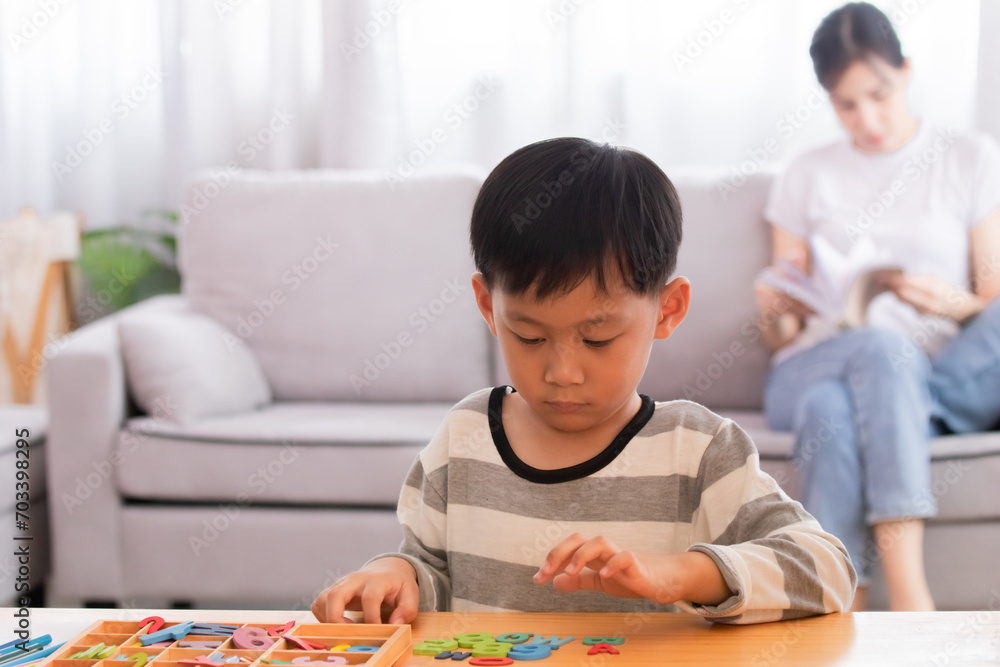 Cute Asian preschool boy has fun enjoys playing colorful toys alone in living room with mother, female mom reading book sitting on sofa relaxing with little kid after homeschool teaching using laptop