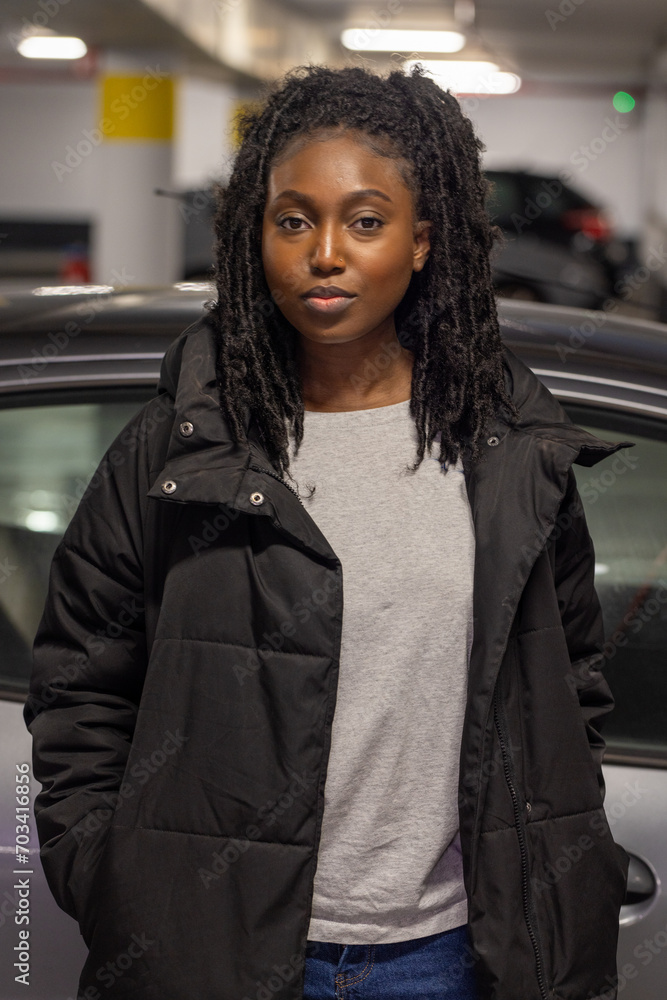 A young Black woman stands poised in an urban parking garage, her style effortlessly blending casual and cool. She wears a timeless black puffer jacket over a classic gray tee, with her natural locs A young Black woman stands poised in an urban parking garage, her style effortlessly blending casual and cool. She wears a timeless black puffer jacket over a classic gray tee, with her natural locs