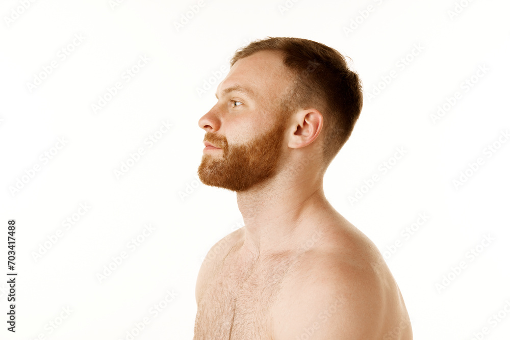Fototapeta premium Side view portrait of young handsome redhead man with beard and bare shoulders looking away against white studio background. Concept of natural beauty, youth, spa treatment, selfcare, cosmetic. Ad
