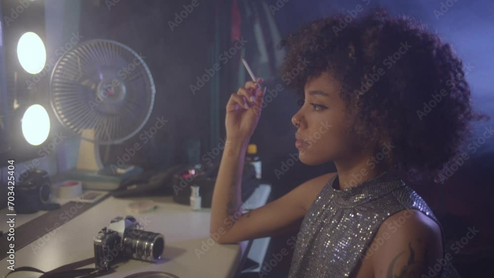 Afroamerican young woman tasting a cigarette in front of an old fashioned mirror, wearing a glitter dress.