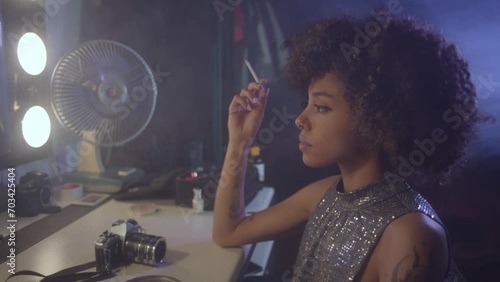 Afroamerican young woman tasting a cigarette in front of an old fashioned mirror, wearing a glitter dress.