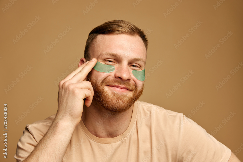 Fototapeta premium Portrait of overjoyed young man with beard applying hydrogel patches with moisturized effect under eyes against beige background. Concept of beauty treatment, selfcare and hygiene, cosmetic, spa.