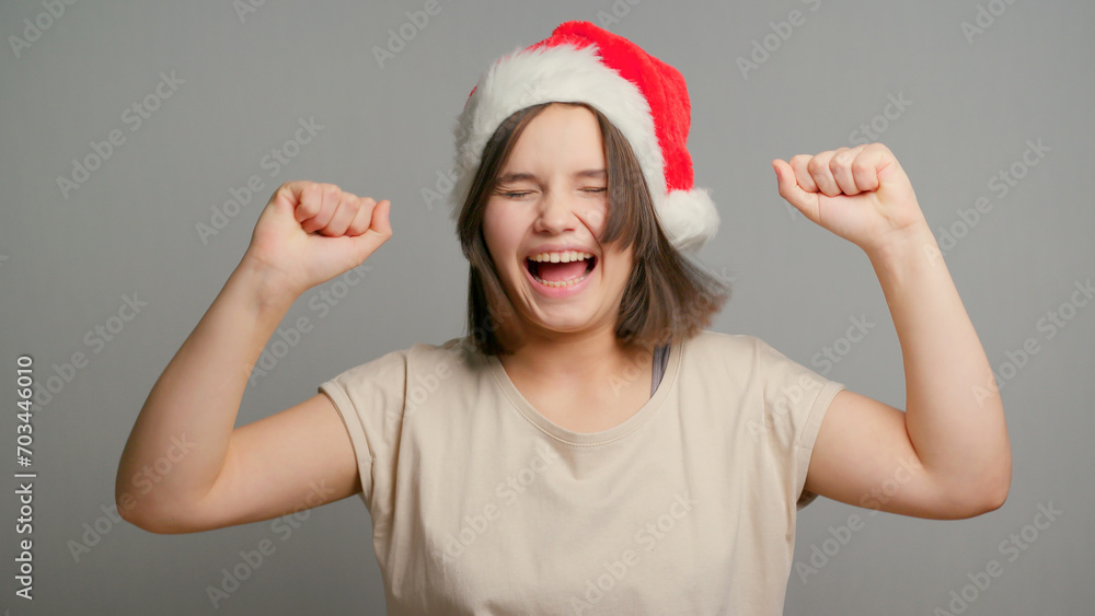 Young latin teenager female student in red santa hat happy dancing celebrating new year isolated on gray background. Copy space Young latin teenager female student in red santa hat happy dancing celebrating new year isolated on gray background. Copy space