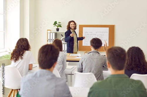 Young male teacher standing in front high school students sitting back at the desks in classroom during a lesson. Boy student raising hand to answer a question. Education and teaching concept. photo