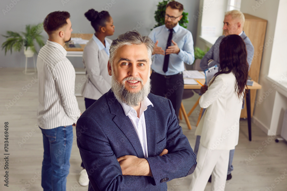 Portrait of senior businessman at work. Happy mature bearded business man in suit looking at camera while standing in office, with team of diverse employees or colleagues in background Portrait of senior businessman at work. Happy mature bearded business man in suit looking at camera while standing in office, with team of diverse employees or colleagues in background