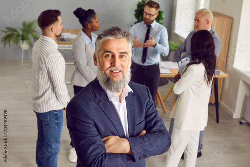 Portrait of senior businessman at work. Happy mature bearded business man in suit looking at camera while standing in office, with team of diverse employees or colleagues in background