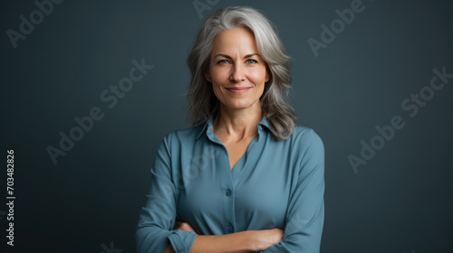 Mature woman with long gray hair, wearing a blue shirt, smiling at the camera with her arms crossed against a blue-gray background.