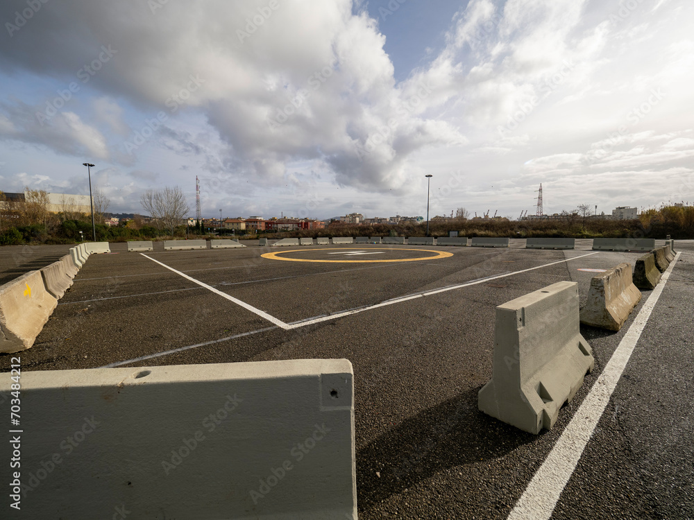 A closeup of an asphalt-covered helipad with a special symbol in the center for helicopter landing, against the backdrop of a green field and a cloudy evening sky. A closeup of an asphalt-covered helipad with a special symbol in the center for helicopter landing, against the backdrop of a green field and a cloudy evening sky.