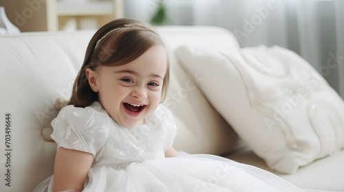 A smiling little girl with Down syndrome in light clothes is sitting on the couch. Down's day.