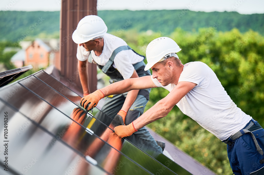 Workers building photovoltaic solar panel system on rooftop of house. Men technicians in helmets and gloves installing solar module with help of hex key outdoors. Alternative energy. - obrazy, fototapety, plakaty 