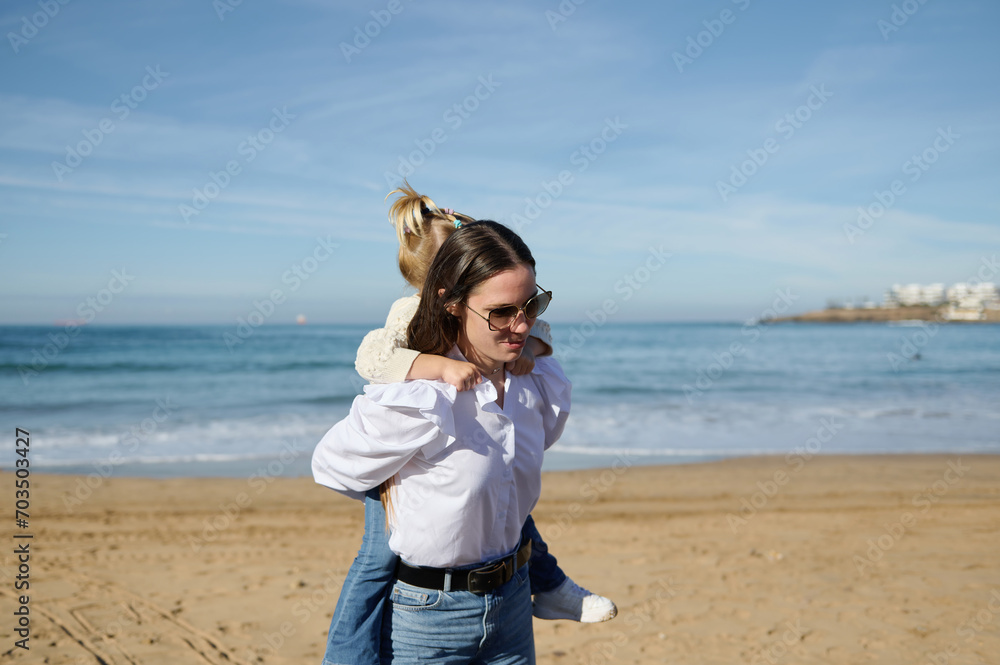 Young mother carries her little daughter on her back, walking along the beautiful waves breaking on the Atlantic shore Young mother carries her little daughter on her back, walking along the beautiful waves breaking on the Atlantic shore