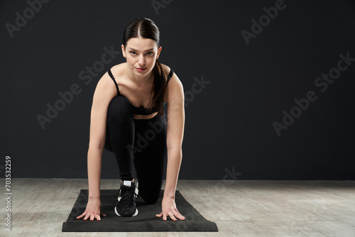 Athletic sporty woman in activewear standing in crouch start pose and looking at camera. Front view of caucasian sportswoman kneeling, while exercising on yoga mat indoors. Concept of sport, training.
