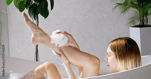 Focused girl washing neat legs, chilling in comfortable bathtub in luxury hotel. Side view of beautiful caucasian female scrubbing body with shower sponge, in front of grey wall. Self care concept.