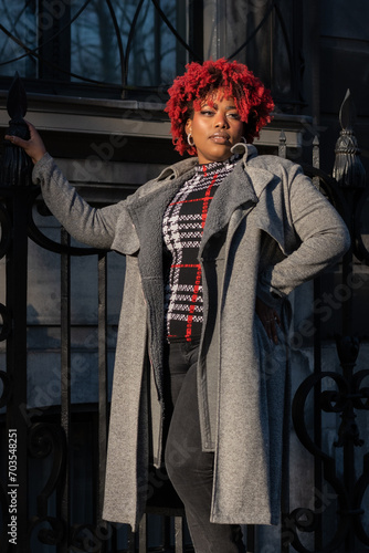 Female model with strong unique style appearance in urban wealthy inner city street town house iron fence environment. Confident curvy black lady stylish red hair with winter good taste fashion 