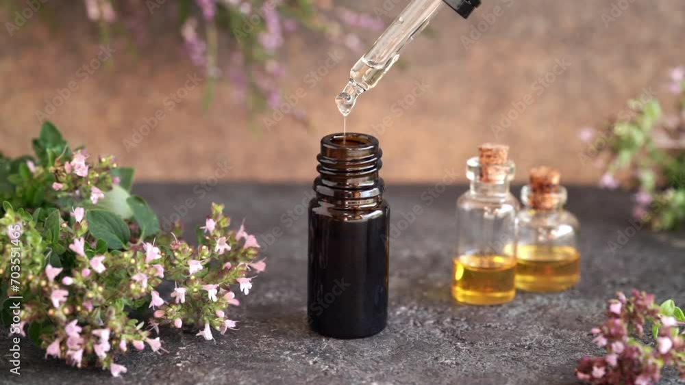 Drops of aromatherapy essential oil are falling from a dropper into a brown bottle, with fresh oregano flowers