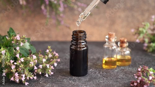 Drops of aromatherapy essential oil are falling from a dropper into a brown bottle, with fresh oregano flowers