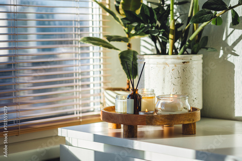 Apartment decoration with aroma reed diffuser, burning aromatic candles on wooden tray. Hygge, home aromatherapy, relaxation concept. Green plants on background. Bali oriental style in modern interior photo