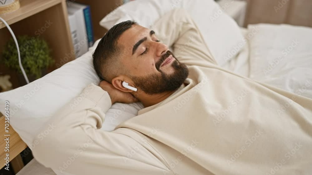 A bearded young man relaxes in bed with wireless earbuds, eyes closed, in a cozy home interior, exhibiting tranquility and comfort. A bearded young man relaxes in bed with wireless earbuds, eyes closed, in a cozy home interior, exhibiting tranquility and comfort.