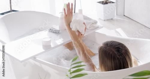 Relaxed woman with short hair washing arms, while taking bath at home, in motion. View from above of anonymous bare female taking bath, using tray with bath accessories. Concept of spa, self-care.