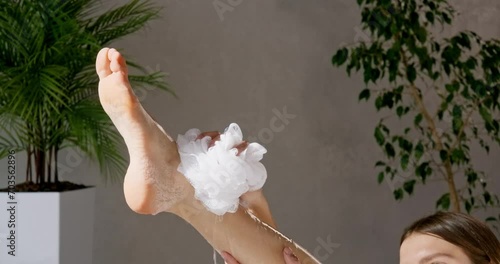 Gorgeous young woman washing body seductively, while lying in comfortable tub. Crop view of calm girl with haircut scrubbing leg, while taking bath, surrounded by plants, in motion. Body care concept.