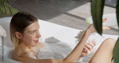 Young blonde woman pouring body wash on sponge, while sitting in bathtub. Side view of seductive female scrubbing legs with sponge, while taking bath at home. Self care, hygiene, cosmetics concept.