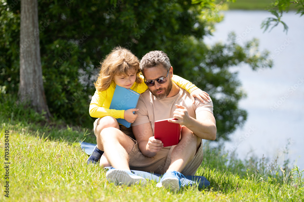 Father reading book with son in park, preparing school homework together, parenting. Summer lifestyle. Parenting and childhood concept. Little boy learning with father in outdoors garden. Father reading book with son in park, preparing school homework together, parenting. Summer lifestyle. Parenting and childhood concept. Little boy learning with father in outdoors garden.