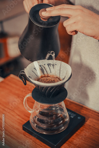 A person pouring hot water from a black kettle into a coffee dripper on top of a clear glass carafe on a table