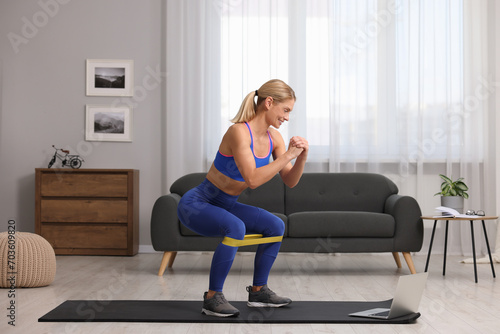Fit woman doing squats with fitness elastic band near laptop on mat at home