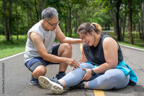 Overweight young woman with knee pain sitting on the running track at local park with both hands grabbing on her trouble knee while a middle age man kneeling beside her helping with concern