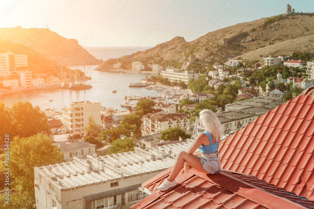 Woman sits on rooftop, enjoys town view and sea mountains. Peaceful rooftop relaxation. Below her, there is a town with several boats visible in the water. Rooftop vantage point.